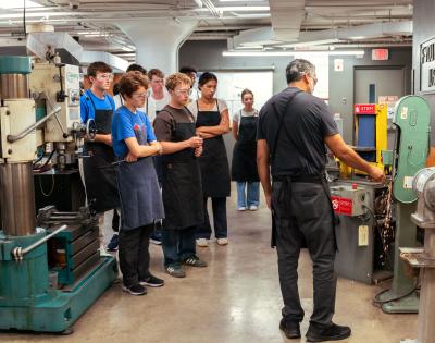 A group of students wearing safety goggles and black aprons attentively observes an instructor operating a machine.