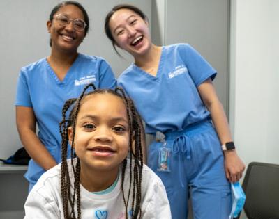 A young girl with braids in a white shirt poses in front of two dental students.