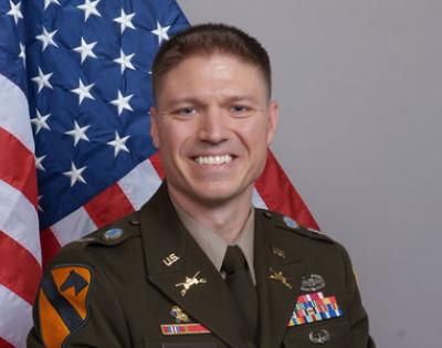 Person wearing military uniform, smiling in front of American flag