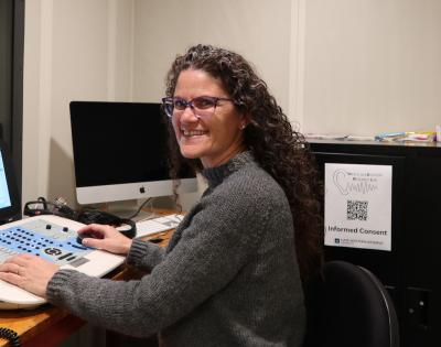 A woman sits with hearing assessment equipment in a sound-isolated booth