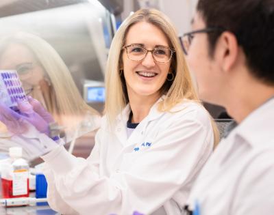 A woman in a lab coat and gloves smiles while holding a test tube rack, engaging with a colleague.
