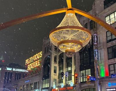 Snow falls on an illuminated street with a large, ornate chandelier arch over the road. 