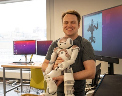 Austin holds a robot in front of a large wall-mounted screen displaying a robot silhouette