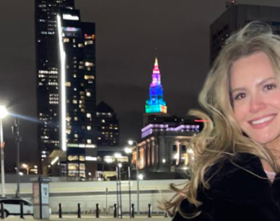 Caroline Deyoe in black coat stands smiling in front of the Cleveland skyline lit up at night.