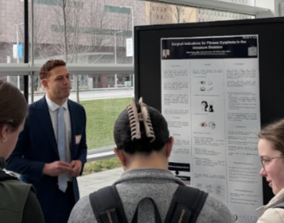 Students gather around a poster presentation being given by a man in a black suit jacket in front of a glass wall.