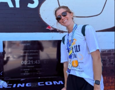 Elizabeth Ginsburg in sunglasses and wearing a race medal stands in front of a computer screen displaying her race time.