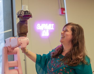 A scientist smiles and looks at a pink robotic arm with a sock puppet