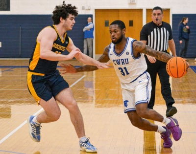 A basketball player in a white CWRU jersey dribbles the ball past a defender in a navy and yellow jersey. A referee observes in the background. 