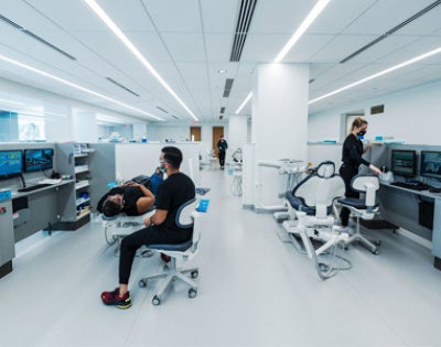 A modern dental clinic with sleek white interiors and several patients receiving care. Dentists and staff, wearing black uniforms and masks, attend to patients in dental chairs.