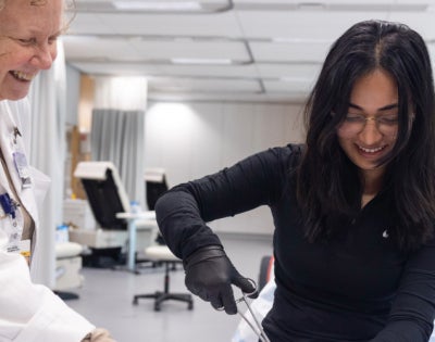 A student in a black shirt practice sutures while a professor in a white lab coat watches and smiles.