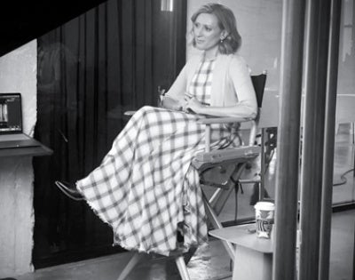 Black and white image of a woman seated in a director's chair, wearing a long checkered dress. She appears calm and focused, with a coffee cup on a side table.