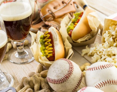 Spread featuring hot dogs, popcorn, peanuts, two glasses of beer, baseballs, a glove, and a bottle opener on a wooden surface.