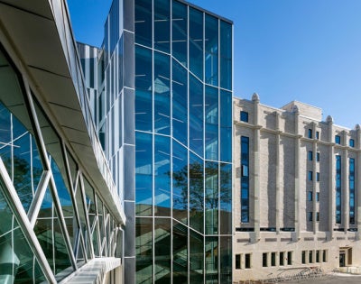 Modern glass building with angular design adjacent to a classic stone structure under clear blue sky, conveying contrast between contemporary and traditional architecture.