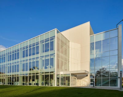 A modern, glass-fronted building with three stories reflecting clear blue skies, set against a backdrop of trees. The scene conveys a crisp, professional atmosphere.
