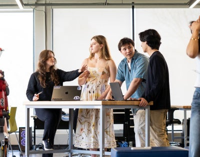 A group of Case Western Reserve University students stand in a classroom, some around a table; robotics are in the corner