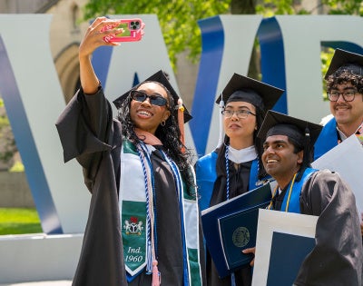 Four graduates in their caps and gowns posed for a selfie in front of the CWRU letters