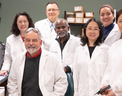Seven researchers in white lab coats pose together inside a biomedical laboratory. Shelves lined with binders and lab supplies surround the group, while a computer monitor on the left displays colorful microscopic cell images. 