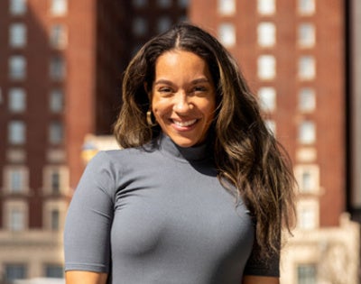 Smiling woman in a gray dress stands outside with tall red brick building in the background. Bright sunlight creates a warm and cheerful atmosphere.