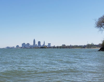 A calm lake with gentle waves is in front of the Cleveland skyline, visible in the distance. A cluster of trees stands on the right shoreline.