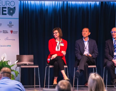Panel of five professionals seated on stage at NeuroNEO 2026 conference. The speaker stands at a podium with banners and plants nearby.