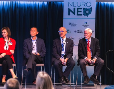 Panel of five professionals seated on stage at NeuroNEO 2026 conference. The speaker stands at a podium with banners and plants nearby.