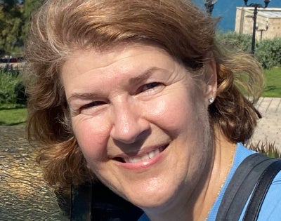 A smiling woman with short brown hair wearing a blue shirt and necklace, outdoors on a sunny day with trees and lampposts in the background.