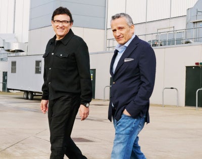 Two men are walking on a concrete surface outside an industrial building. One wears a black jacket, the other a navy blazer and jeans. Both are smiling.