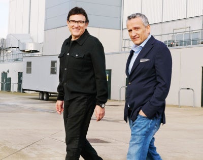 Two men are walking on a concrete surface outside an industrial building. One wears a black jacket, the other a navy blazer and jeans. Both are smiling.