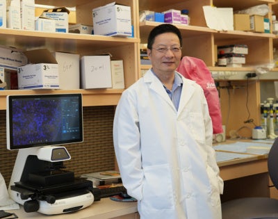 Zhenghe “John” Wang, in a lab coat, stands next to a screen showing a fluorescence image of tumor tissue. In the background are shelves filled with laboratory equipment. 