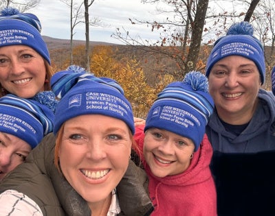 Group of six smiling people in blue beanies from Case Western Reserve University School of Nursing. They're outdoors with a scenic, wooded background.