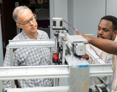 Two men, focused and engaged, work together on a complex machine in a lab. One points attentively while the other observes, discussing adjustments.