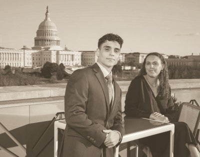 Felipe Joglar-Viera and Shannon Moore in formal attire sit at an outdoor table, with the U.S. Capitol in the background. 