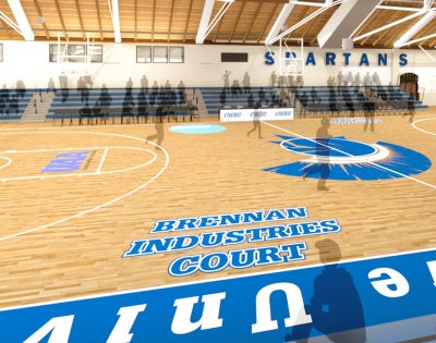 Interior of a gymnasium featuring a basketball court labeled "Brennan Industries Court" with blue and white bleachers and "Spartans" on the wall.