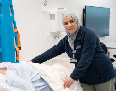 A nursing professor is placing a blanket on a pregnant, high-fidelity mannequin lying on a bed. The background mimics the hospital setting. 