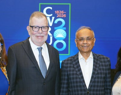 Four people stand smiling against a blue backdrop with "CV 20-26" text. They are dressed in formal attire, reflecting a professional event.