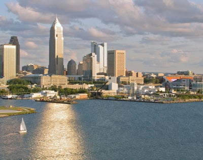 Photo of the Cleveland skyline on a summer day, with the sun reflecting on Lake Erie and a sailboat on the water