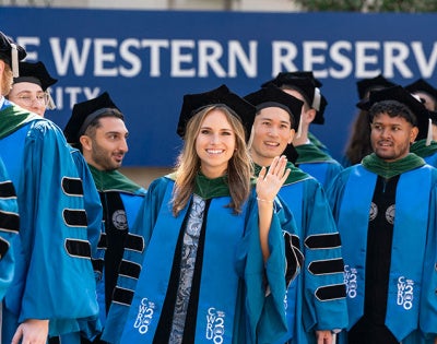 A CWRU grad waves as she walks with a procession to a commencement ceremony and passes the CWRU sign on Adelbert Road and Euclid Avenue