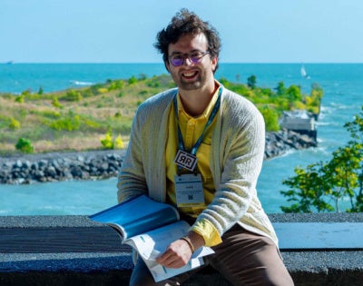 Photo of Sebastian Correa holding a book while posing with a tropical area behind him
