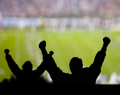 Photo of the silhouettes of fans cheering with a soccer stadium field in front of them