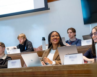 Seven students sit in a tiered classroom in the Peter B. Lewis Building. Two students smile and interact with each other while the others focus on their laptops. 