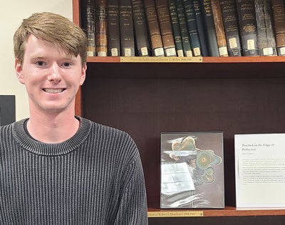 Aidan Selkirk poses for a photo with a copy of his artwork created with an aerosol jet printer framed on a shelf next to him