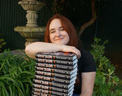 Julia Bianco sitting in the grass with a stack of her new books in her lap