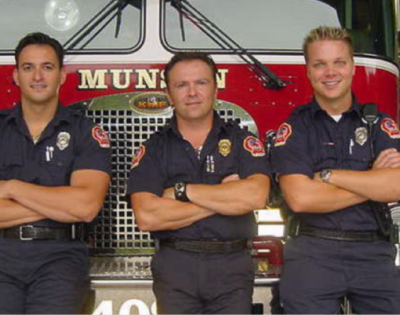 Three men stand in front of a fire truck with arms crossed.