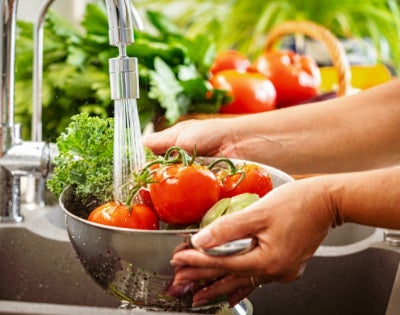 Hands holding a colander full of fresh vegetables under running water in kitchen sink