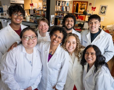 Seven students and a faculty member pose together in white lab coats inside a research laboratory, smiling at the camera. Shelves, equipment, and research materials are visible in the background.