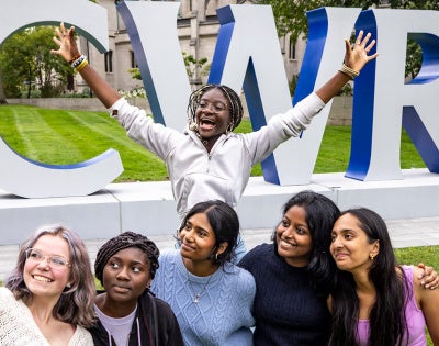 Five CWRU students pose for a selfie photo as another student leaps with their arms up behind them in front of the CWRU letters on campus