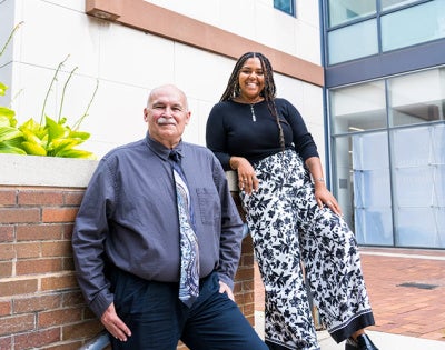 Ric Kruszynski sits on a brick planter wall beside Michaela Jackson-Smith outside a modern glass-and-brick building.