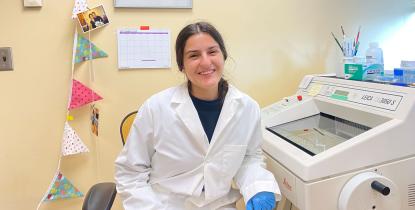 A woman wearing a lab coat and blue gloves smiling in a lab. She is next to scientific equipment.