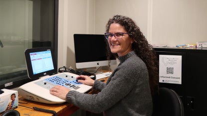 A woman sits with hearing assessment equipment in a sound-isolated booth