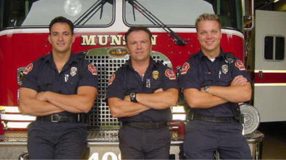 Three men stand in front of a fire truck with arms crossed.